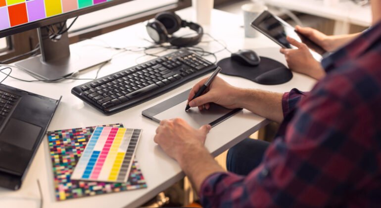 Designer working on a new project in an office; web designer drawing on a drawing pad and choosing colors from a color palette. Focus on the tip of the pen and the fingers