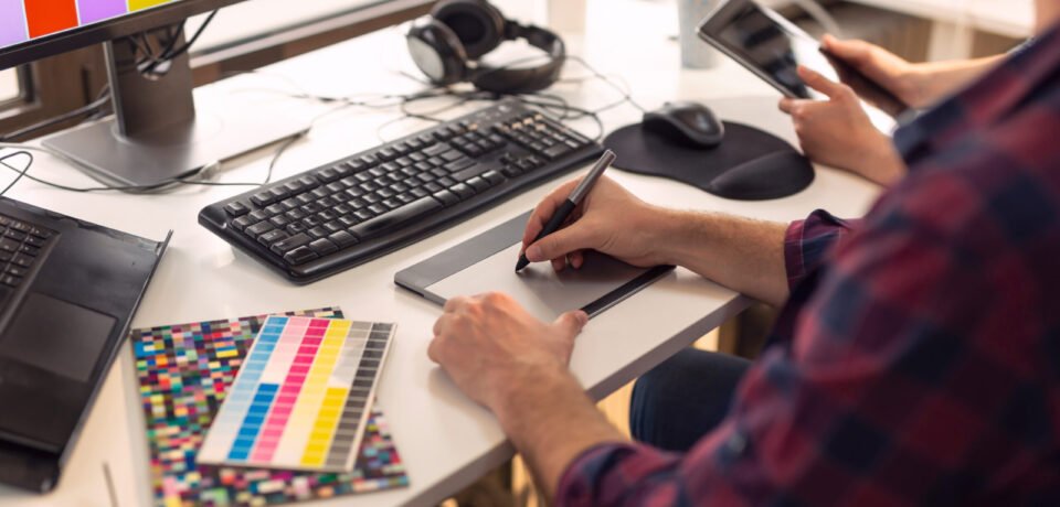 Designer working on a new project in an office; web designer drawing on a drawing pad and choosing colors from a color palette. Focus on the tip of the pen and the fingers